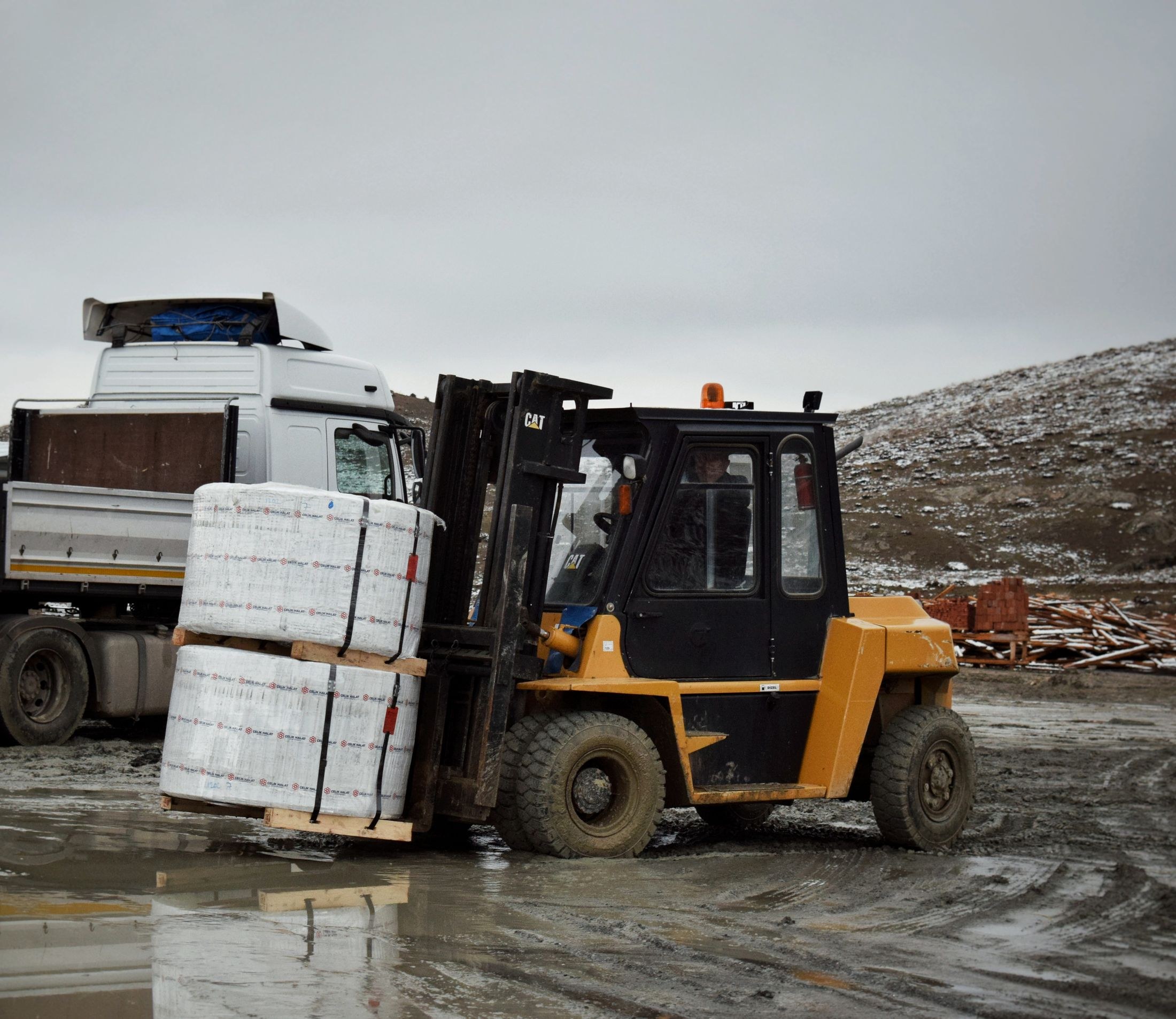 A forklift operator moves two wrapped pallet loads beside a truck in an outdoor industrial yard.